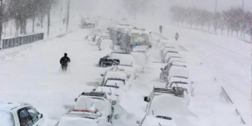 A snowy street with numerous cars buried under snow, some people walking around, and heavy snowfall continuing steadily.