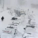 A snowy street with numerous cars buried under snow, some people walking around, and heavy snowfall continuing steadily.