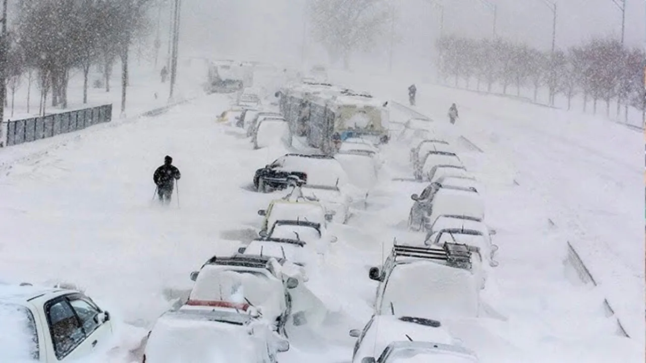 A snowy street with numerous cars buried under snow, some people walking around, and heavy snowfall continuing steadily.