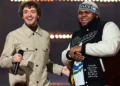 Druski and Jack Harlow stand on stage smiling, one holding a microphone. The man on the left, Druski, wears a beige jacket, while the man on the right, Jack Harlow, sports a black and white jacket with a beanie.