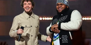 Druski and Jack Harlow stand on stage smiling, one holding a microphone. The man on the left, Druski, wears a beige jacket, while the man on the right, Jack Harlow, sports a black and white jacket with a beanie.