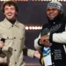 Druski and Jack Harlow stand on stage smiling, one holding a microphone. The man on the left, Druski, wears a beige jacket, while the man on the right, Jack Harlow, sports a black and white jacket with a beanie.