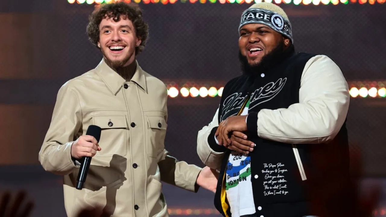 Druski and Jack Harlow stand on stage smiling, one holding a microphone. The man on the left, Druski, wears a beige jacket, while the man on the right, Jack Harlow, sports a black and white jacket with a beanie.