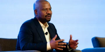 Ted Colbert in a dark suit and white shirt speaks animatedly while seated, gesturing with his hands. With a microphone clip on his shirt, Boeing defense chief Ted Colbert discusses the Starliner program. The background is light blue.
