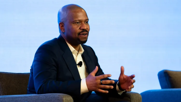 Ted Colbert in a dark suit and white shirt speaks animatedly while seated, gesturing with his hands. With a microphone clip on his shirt, Boeing defense chief Ted Colbert discusses the Starliner program. The background is light blue.