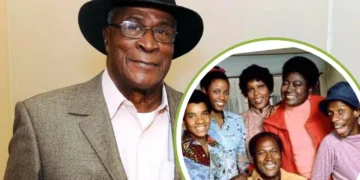 Older man in a hat and suit stands smiling. Inset shows John Amos, the beloved actor, posing and smiling with a group of six people.