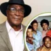 Older man in a hat and suit stands smiling. Inset shows John Amos, the beloved actor, posing and smiling with a group of six people.