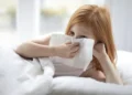 A U.S. child with red hair lies in bed using a tissue to blow their nose, possibly fighting off walking pneumonia as the crisp air of Fall 2024 settles in.