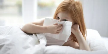A U.S. child with red hair lies in bed using a tissue to blow their nose, possibly fighting off walking pneumonia as the crisp air of Fall 2024 settles in.