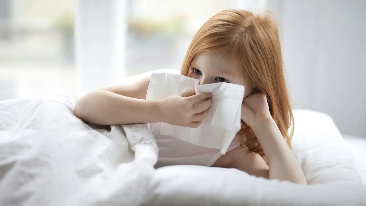 A U.S. child with red hair lies in bed using a tissue to blow their nose, possibly fighting off walking pneumonia as the crisp air of Fall 2024 settles in.