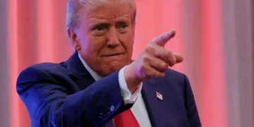 President Trump a suit, reminiscent of a Trump administration appointee, pointing forward as he stands in front of a red and white curtain backdrop.