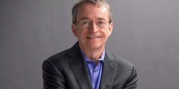 Pat Gelsinger wearing glasses, a dark suit, and a blue shirt smiles confidently against a gray background, embodying the poised demeanor of a CEO from a tech giant.