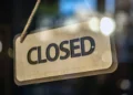 A "Closed" sign hangs on a glass door at Bridgeport Shopping Center, with blurred lights of major retailers shining in the background.
