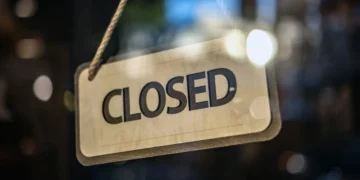 A "Closed" sign hangs on a glass door at Bridgeport Shopping Center, with blurred lights of major retailers shining in the background.
