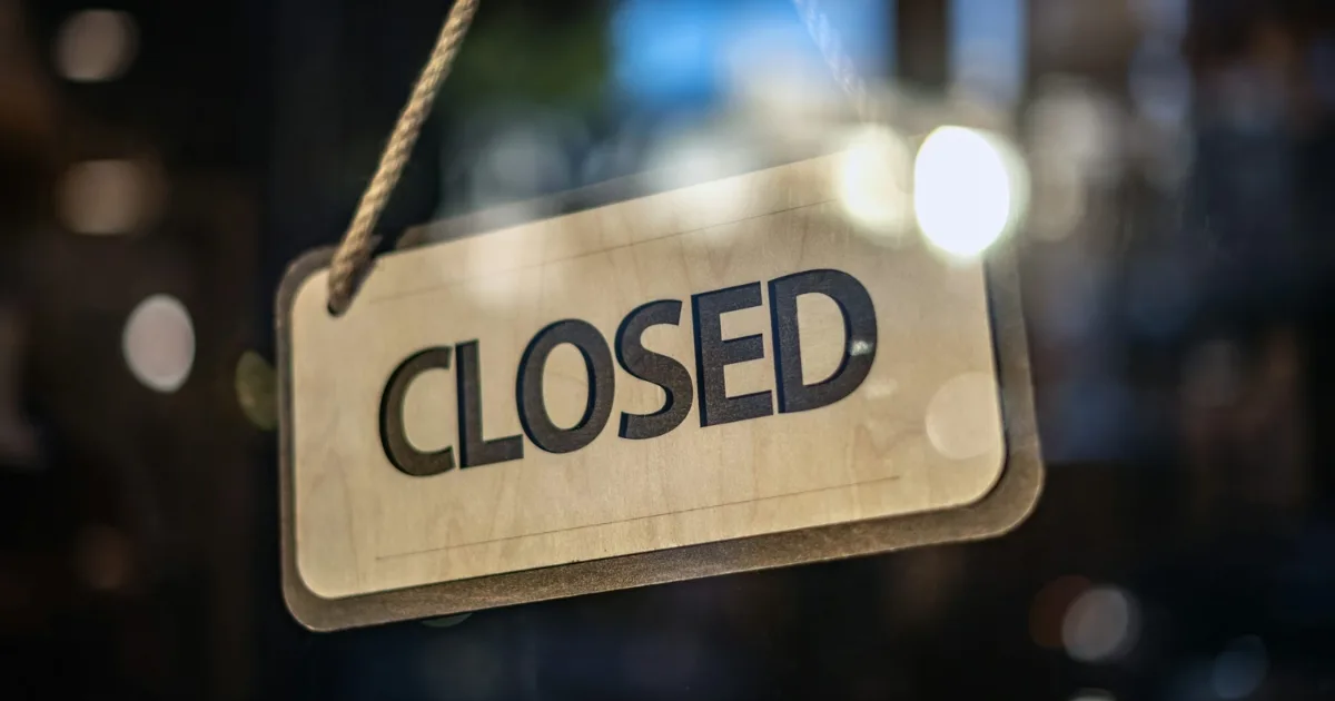 A "Closed" sign hangs on a glass door at Bridgeport Shopping Center, with blurred lights of major retailers shining in the background.