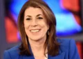 Tammy Bruce, with shoulder-length brown hair, smiles warmly in a blue blazer against a blurred, colorful background reminiscent of official settings.