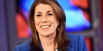 Tammy Bruce, with shoulder-length brown hair, smiles warmly in a blue blazer against a blurred, colorful background reminiscent of official settings.