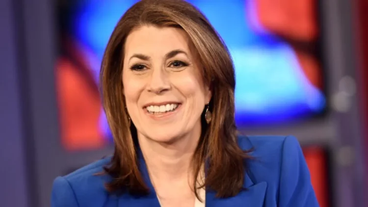 Tammy Bruce, with shoulder-length brown hair, smiles warmly in a blue blazer against a blurred, colorful background reminiscent of official settings.